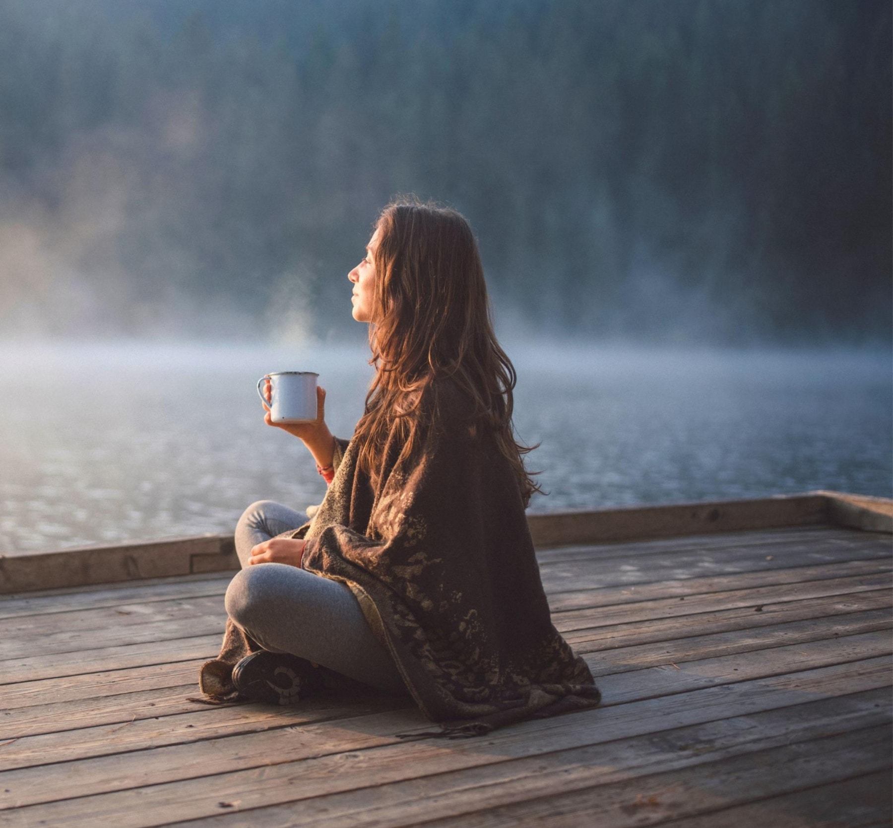 Femme détendue savourant une boisson chaude face à un lac au lever du jour — moment de sérénité et de pleine conscience.