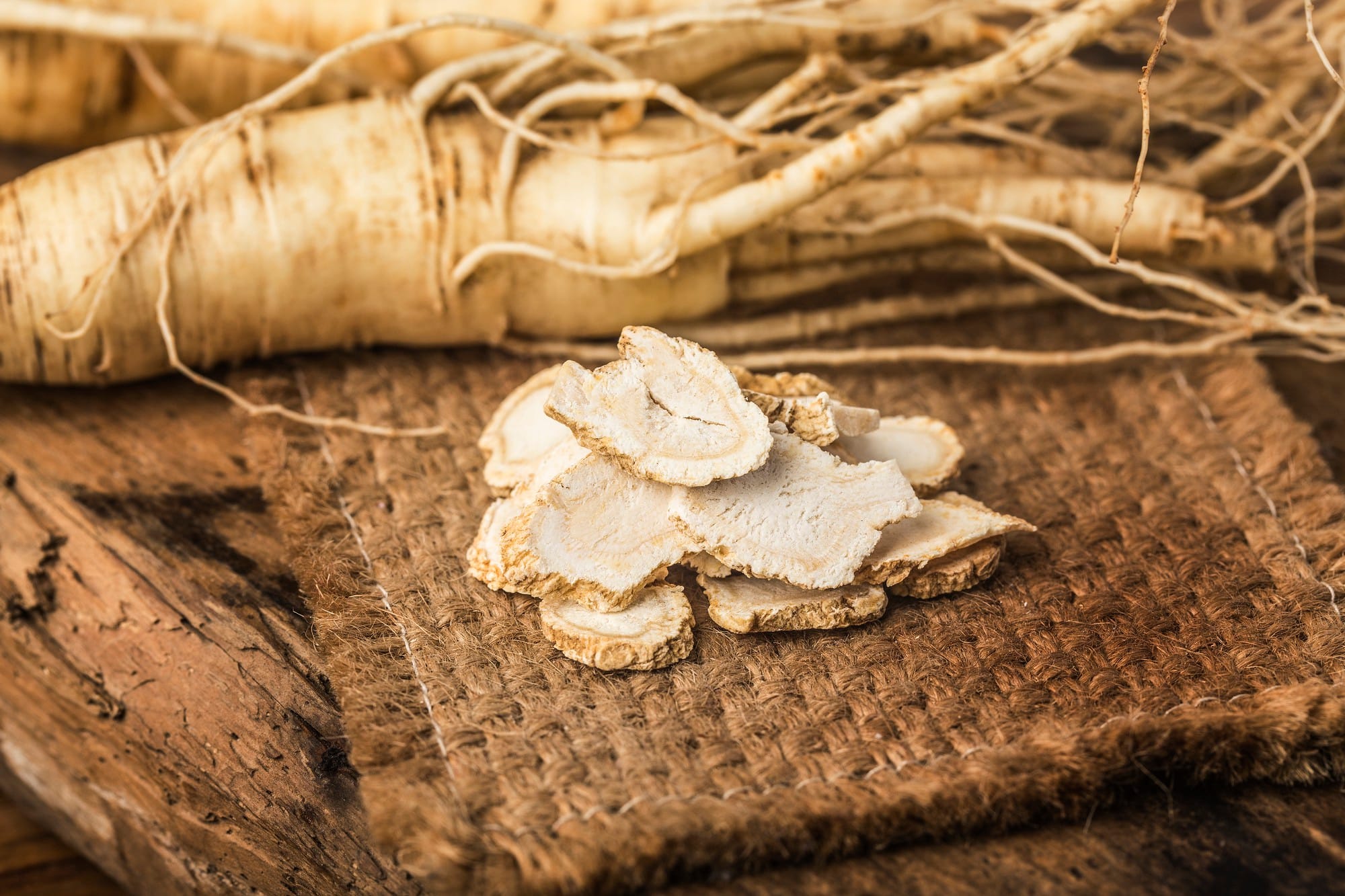 Tranches de racine de ginseng séchée devant des racines entières sur un support en fibres naturelles.