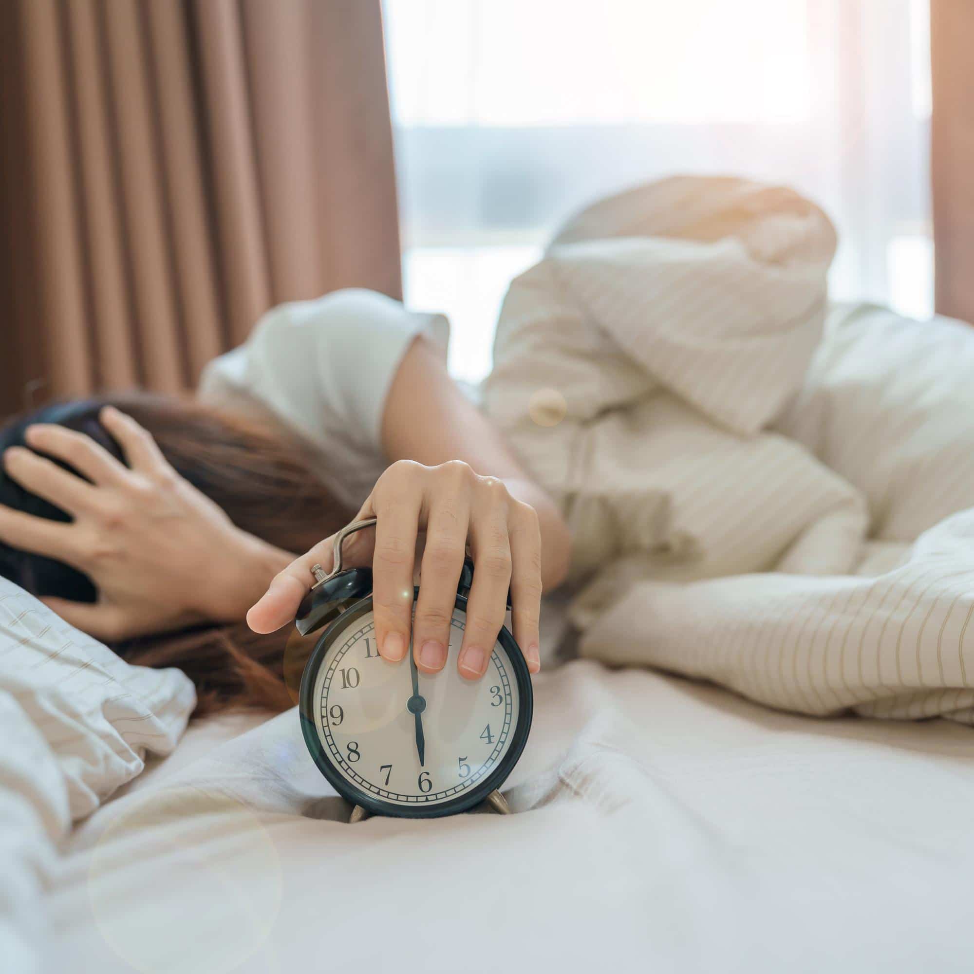 Main éteignant un réveil tandis qu'une personne épuisée reste allongée dans son lit, illustrant le manque de sommeil et son influence sur la libido.