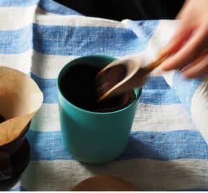 Une main verse du marc de café dans un récipient en céramique bleue, sur une nappe rayée bleue et blanche – préparation d’astuces zéro déchet maison