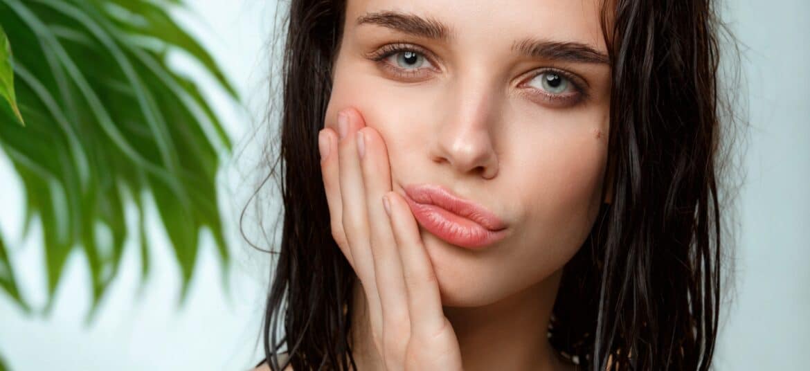Jeune femme au teint frais posant avec une expression pensive, entourée de feuilles vertes sur fond clair – symbole de beauté naturelle et ayurvédique.