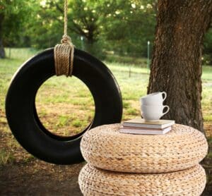 Pouf fait maison en corde naturelle à partir d’un pneu recyclé, posé sous un arbre avec un pneu suspendu en arrière-plan