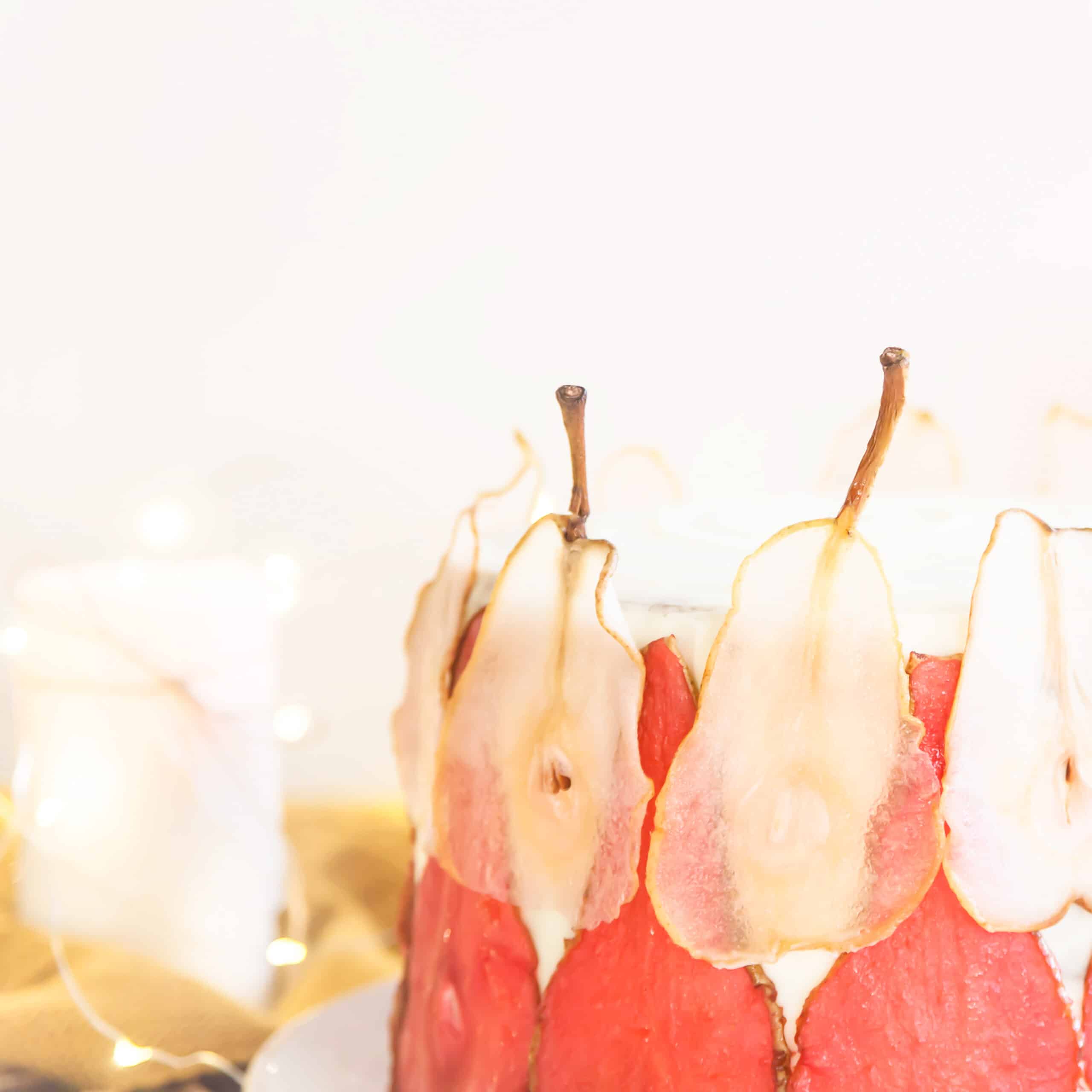 Gâteau de fête décoré de tranches fines de poires rouges et blanches séchées, mis en valeur par une lumière douce et des guirlandes lumineuses en arrière-plan – une idée gourmande et poétique pour revaloriser des fruits trop mûrs.