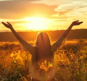 Femme bras levés dans un champ au coucher de soleil, respirant l'énergie estivale.