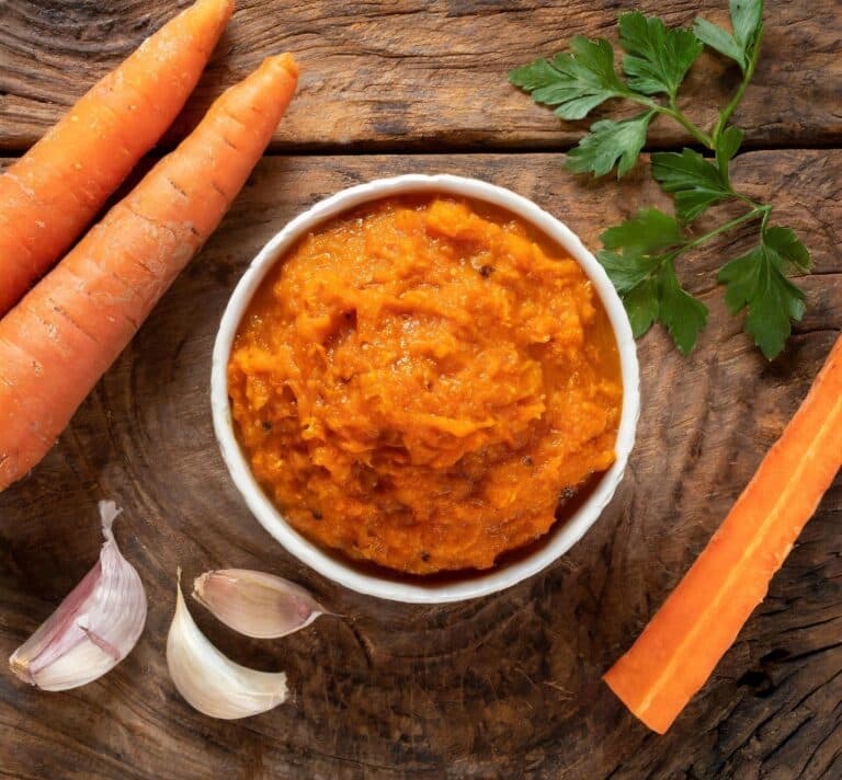 Bol de chutney de carottes au curry fait maison, entouré de carottes et d’ail sur une planche en bois