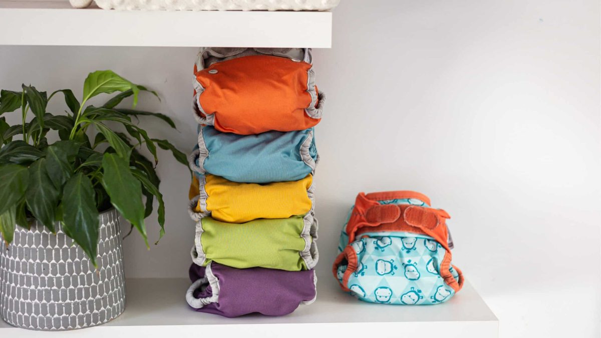 Pile de couches lavables colorées posées sur une étagère à côté d’une plante verte, symbole d’un mode de vie plus écologique et pratique avec bébé.