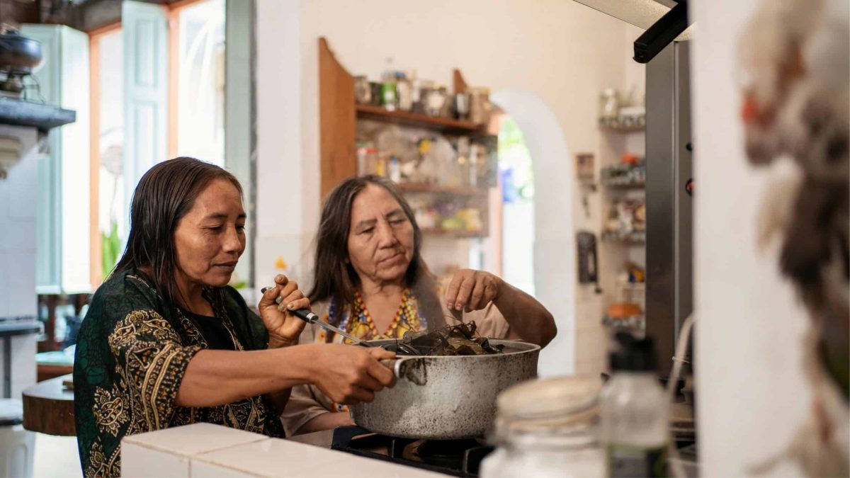 Deux femmes autochtones cuisinant ensemble dans une maison traditionnelle, symbole de solidarité, culture et résistance.