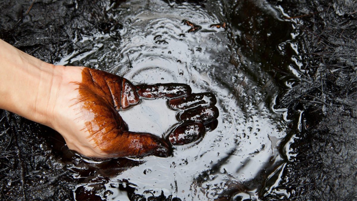 Main humaine recouverte de pétrole brut dans une mare d’hydrocarbures, illustrant la pollution liée aux énergies fossiles.