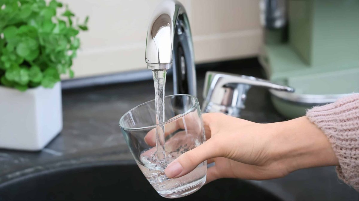 Main remplissant un verre d’eau sous un robinet de cuisine, illustrant la qualité et la composition de l’eau du robinet.