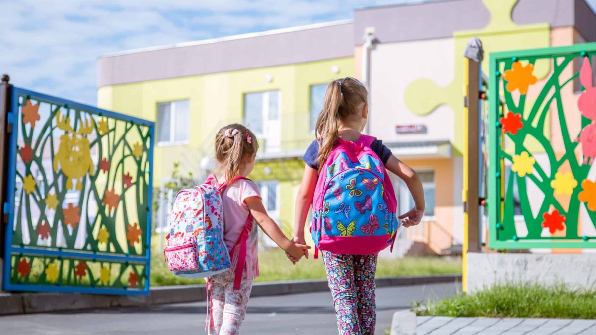 Deux enfants avec cartables entrant à l’école main dans la main, dans une rue apaisée sans voitures.