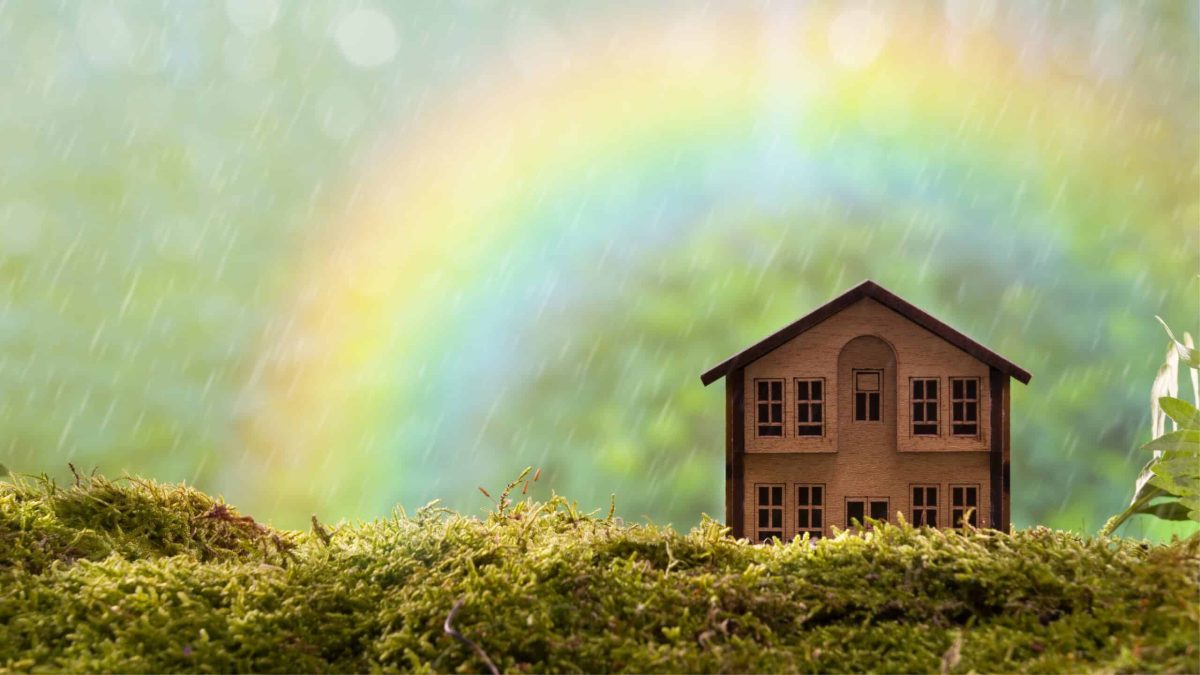 Petite maison en bois posée sur de la mousse verte sous un arc-en-ciel, évoquant un mode de vie écologique et poétique.